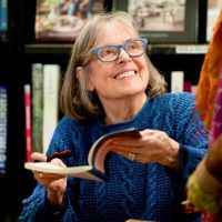 smiling Belgian woman in front of a bookshelf holding an open book and pen and looking up toward someone else