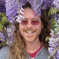 a smiling white man with long hair, light facial hair, and sunglasses surrounded by foliage