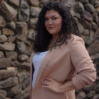 an Ecuadorian-American woman in a blazer standing in front of a wall made of stones with a hand on her hip