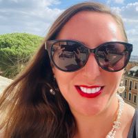 a smiling white woman wearing sunglasses with trees, clouds, and buildings in the background