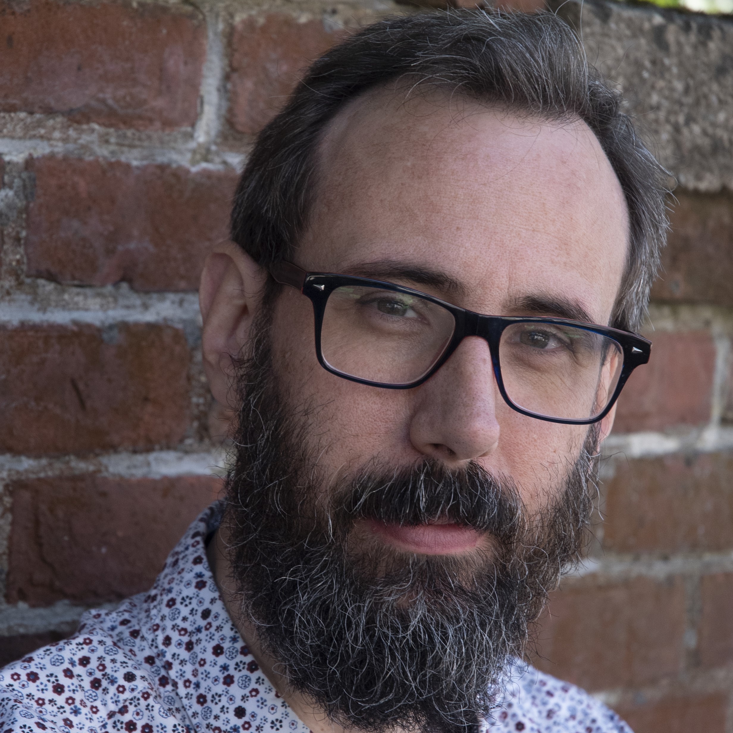 A white man with a beard and glasses in front of a brick wall