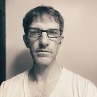 a white man with glasses, short haircut, and a v-neck t-shirt