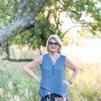 a white woman in sunglasses standing with her hands on her hips in a field with a tree in the background