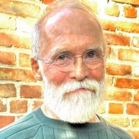 a white man with glasses and a white beard standing in front of a brick wall